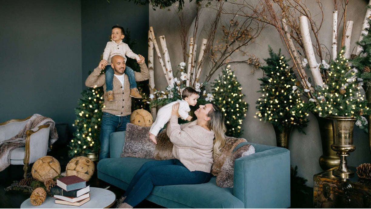 A family of four in a cozy, decorated living room. A woman sits on a teal sofa holding a child, while a man in the background has another child on his shoulders.