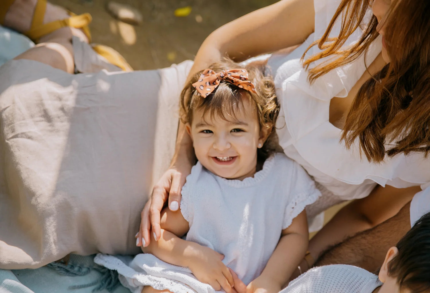 A smiling young child wearing a white dress and a pink bow headband sits on a light-colored blanket outdoors, with an adult in a white top leaning over them in warm natural sunlight.