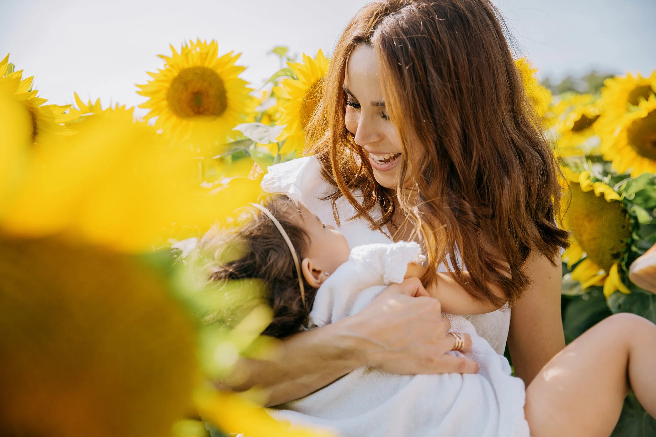 A woman in a white dress holds a baby wrapped in a white blanket while sitting in a field of bright yellow sunflowers.