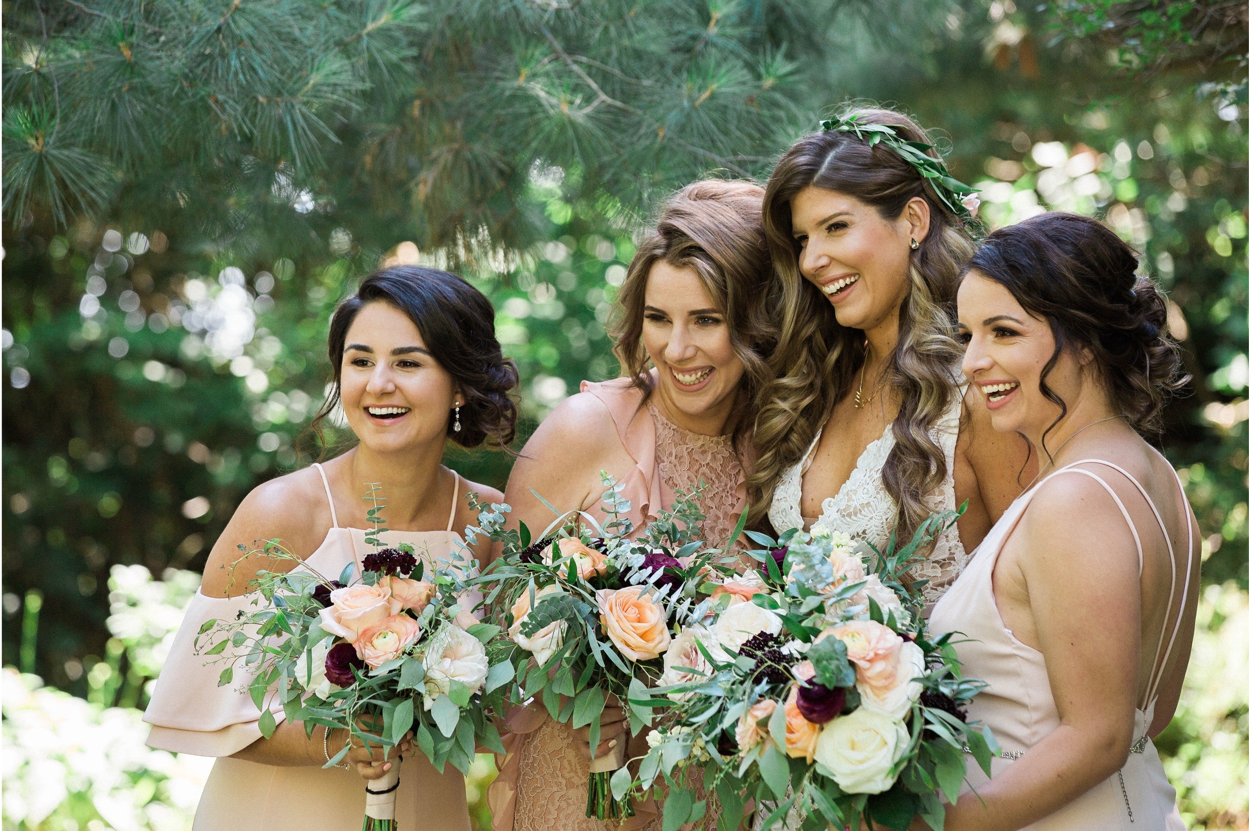 Four bridesmaids laughing together in a forest, one wearing a flower crown