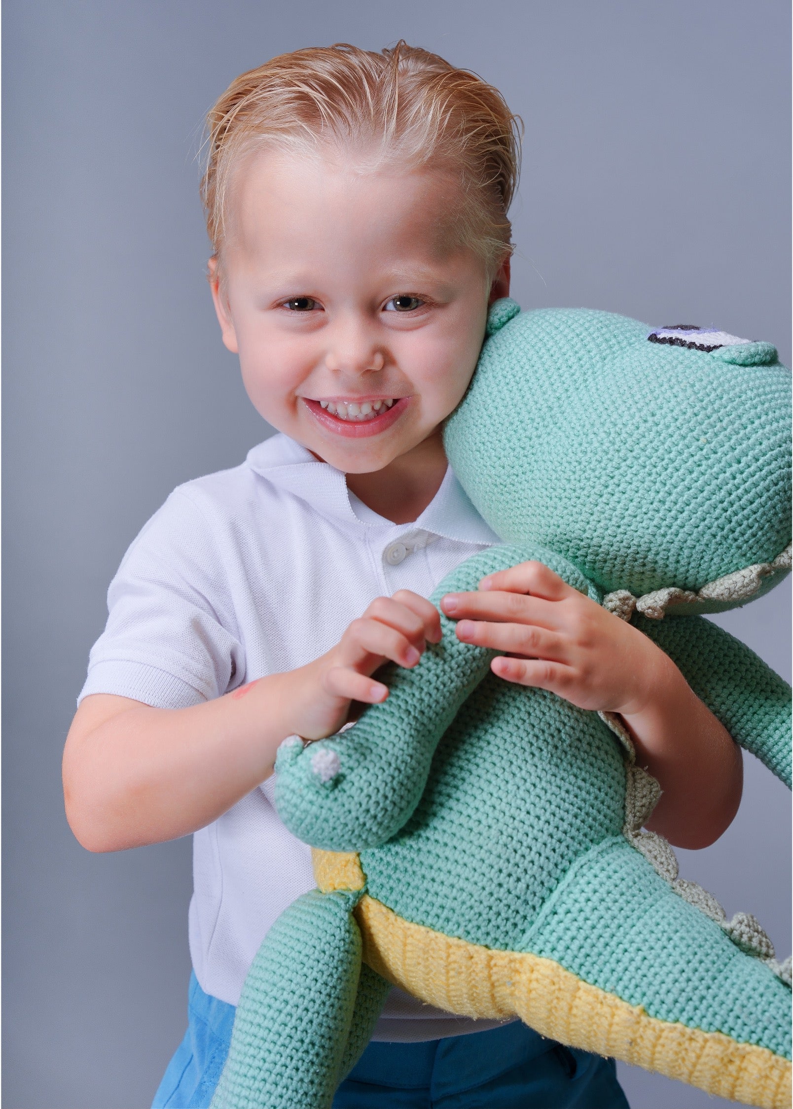 A young blonde boy in a white polo shirt and blue pants smiling while holding a large mint green crocheted dinosaur toy against a solid gray studio background.