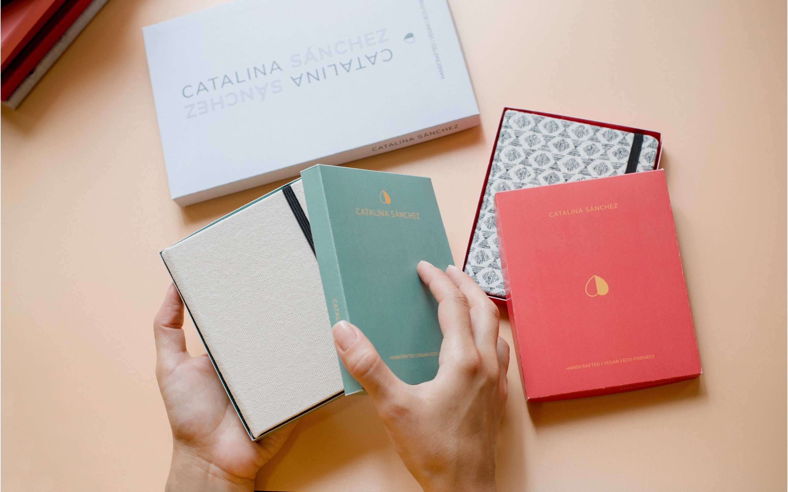 Three hardcover notebooks in beige, mint green, and coral red are held in hands next to their branded white and patterned boxes on a peach-colored surface.