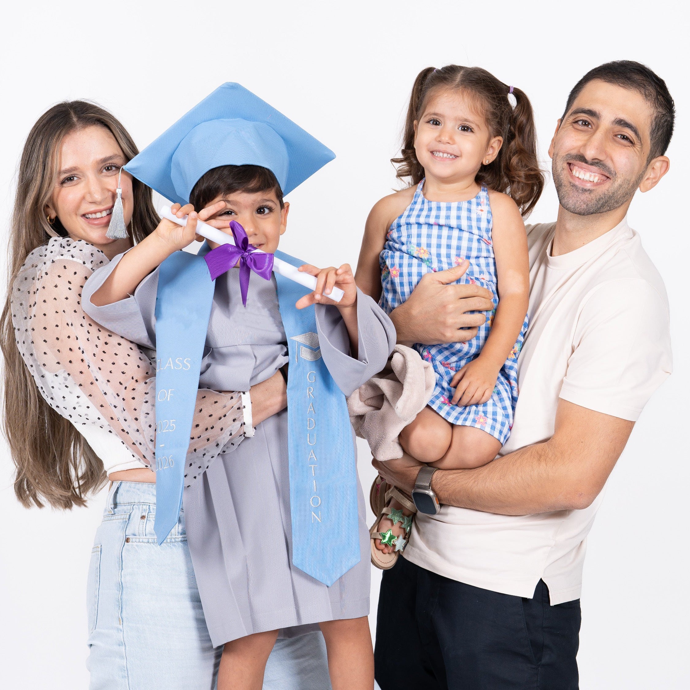 A family of four posing for a photo against a white studio background. The mother holds a young boy wearing a light blue graduation cap and matching gown with a purple ribbon, while the father holds a young girl in a blue and white gingham dress.