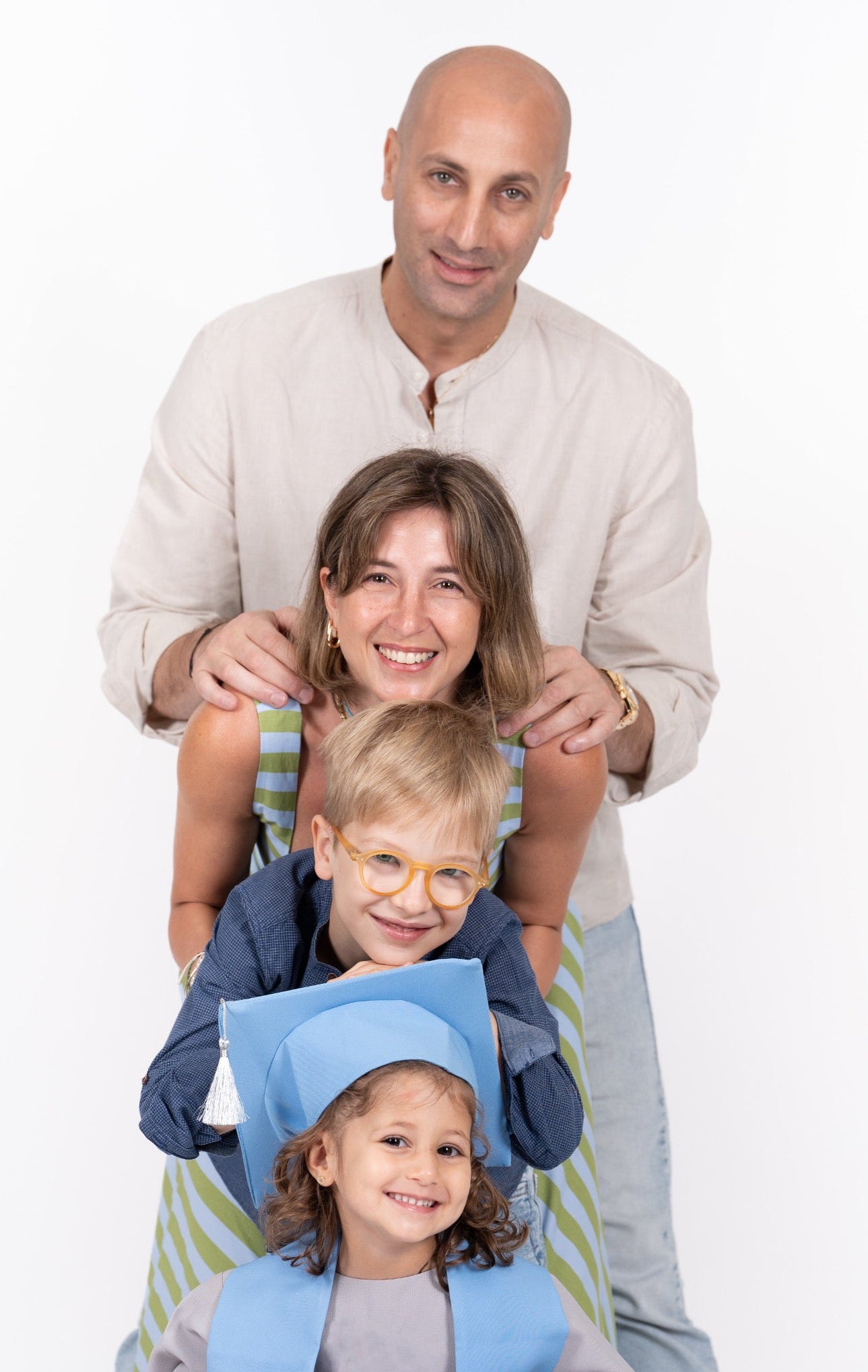 A family of four posing together against a white studio background. The father stands behind the mother, who is holding a young boy wearing glasses and a blue graduation cap and gown.