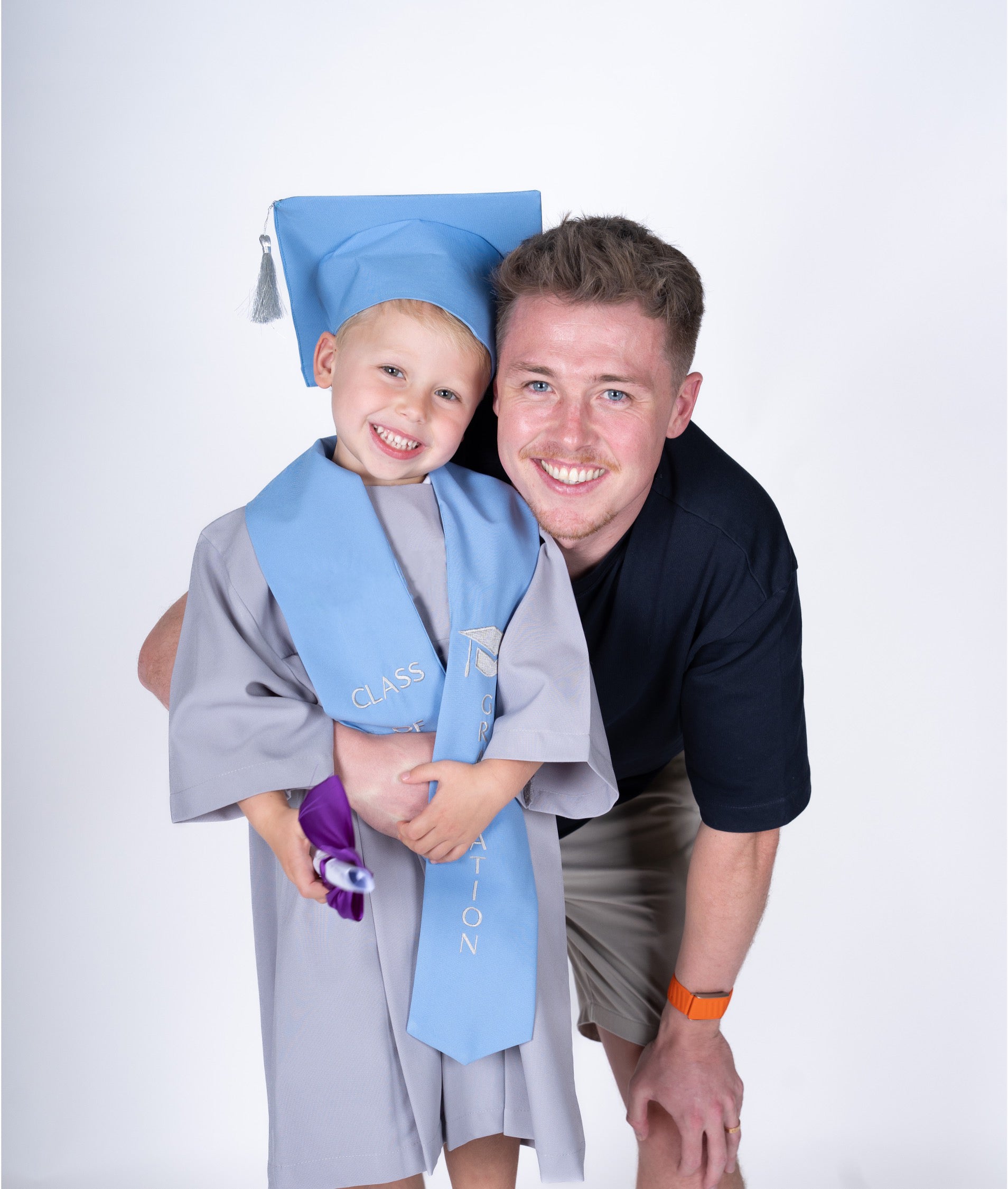 A smiling young boy in a light blue graduation cap and gown stands on his father's shoulders. The father is wearing a dark navy polo shirt and khaki shorts.