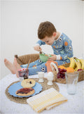 A young boy wearing blue soccer-print pajamas sits on a white tablecloth, eating cereal with a spoon. A basket of fruit, waffles, eggs, and a yellow checkered napkin are arranged around him in a bright, clean indoor setting.