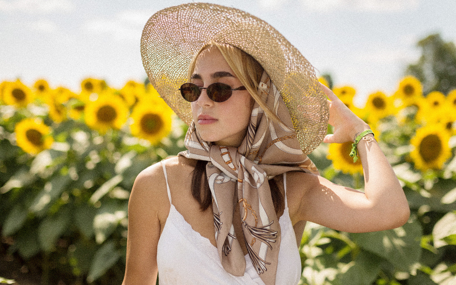 A woman wearing a large straw sun hat, a beige patterned scarf, and sunglasses stands in a sunflower field.