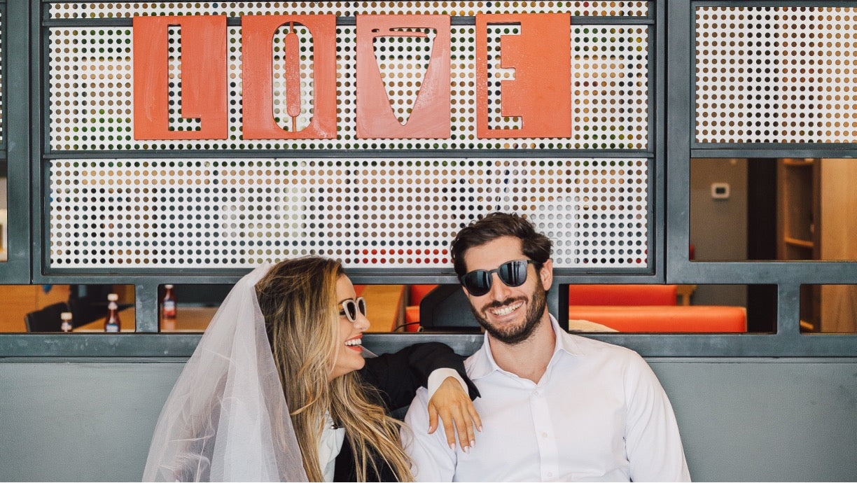 A smiling couple posing together in front of a decorative wall with the word 'LOVE' in orange letters. The woman wears a white wedding veil and sunglasses, and the man wears a white shirt and sunglasses.