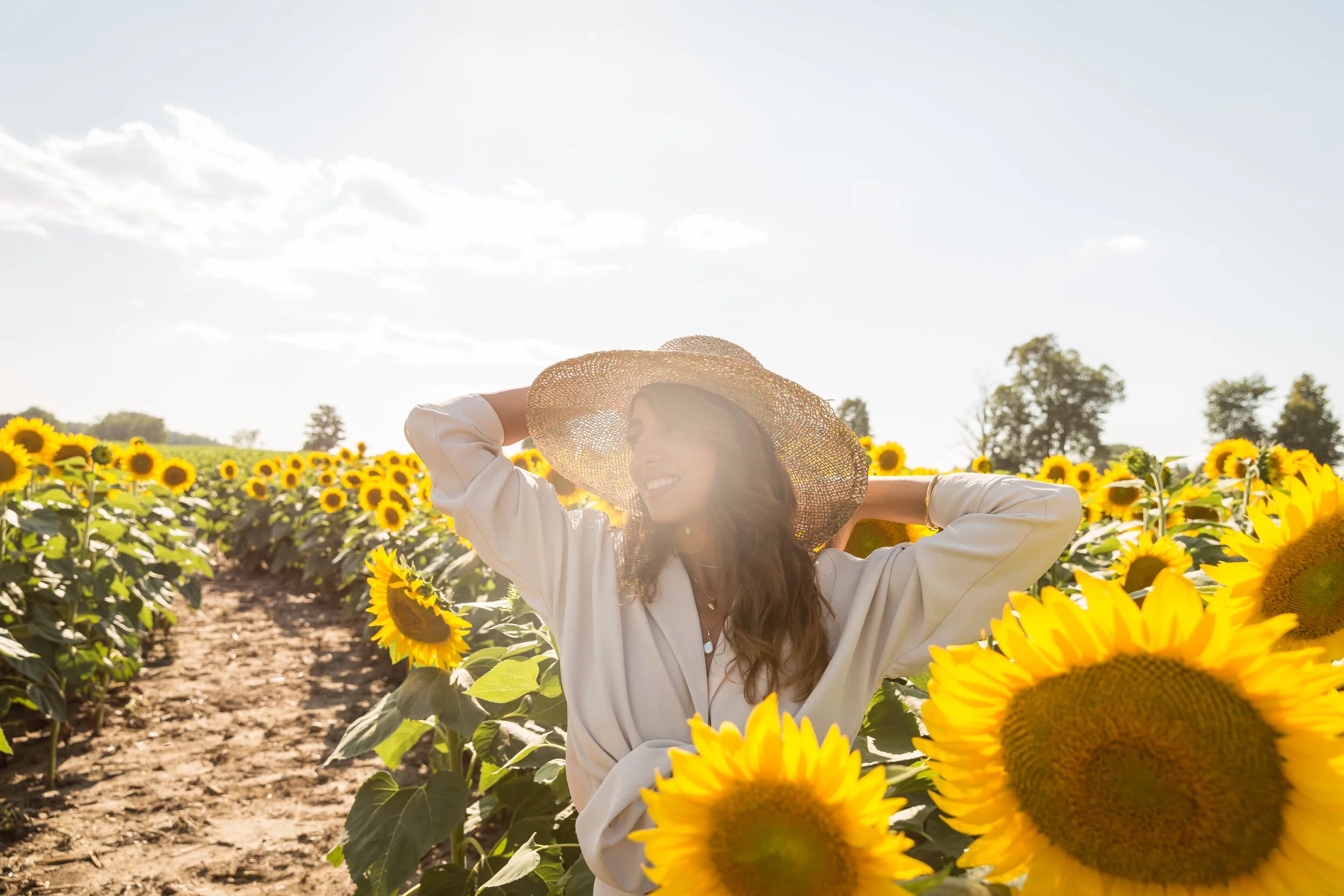 A woman in a white shirt and wide-brim straw hat stands in a sunflower field, smiling in bright sunlight with rows of yellow sunflowers stretching into the background.