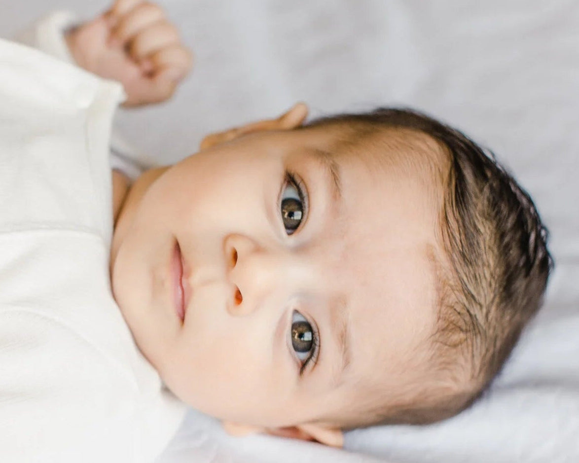A close-up of a baby lying on a white blanket, wearing a white long-sleeve onesie with a small colorful embroidered design on the chest.
