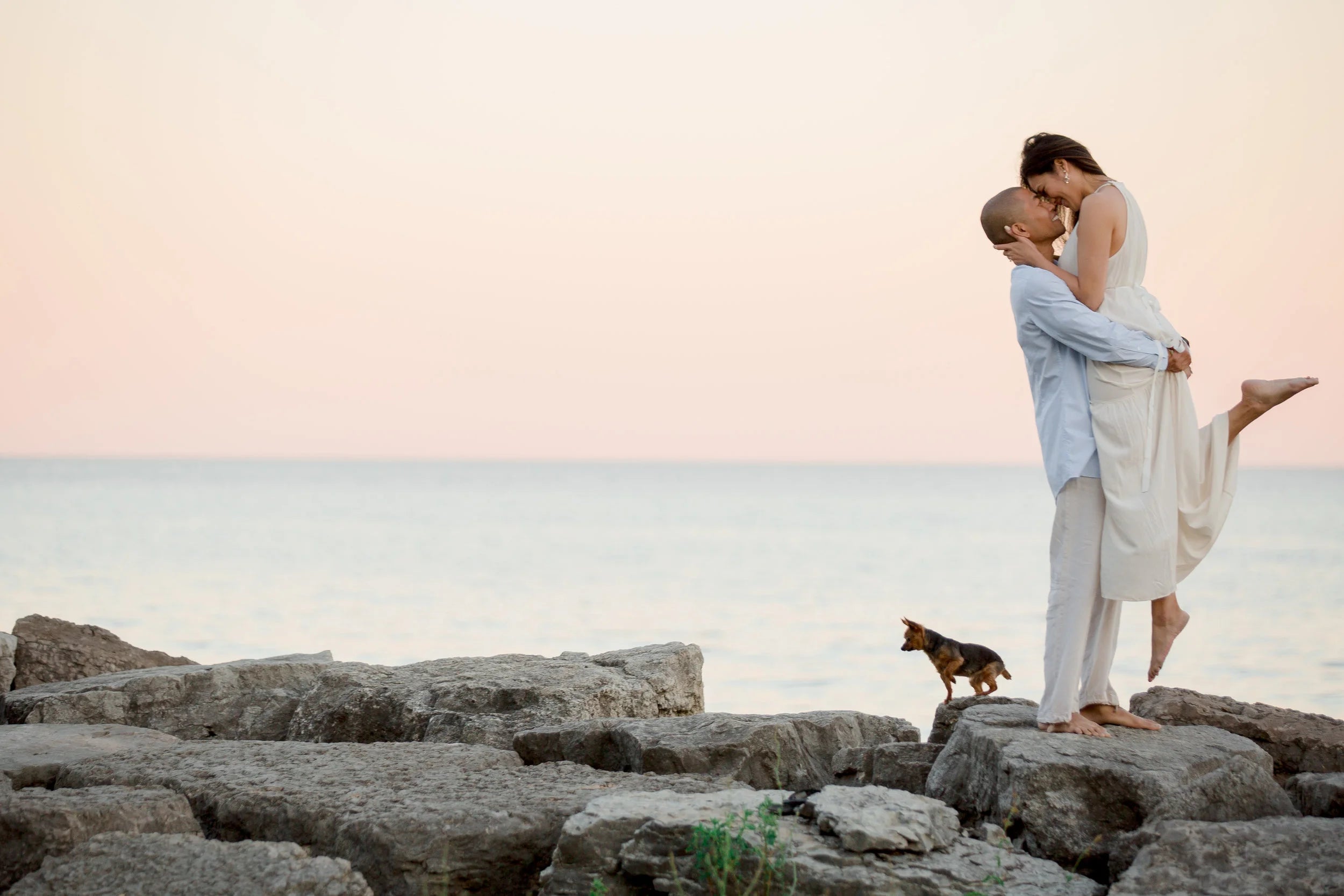 A couple embracing on a rocky shoreline at sunset, with the woman in a flowing white dress and the man in a light blue shirt and white pants. A small dog stands nearby on the rocks, and the ocean and pale sky fill the background.