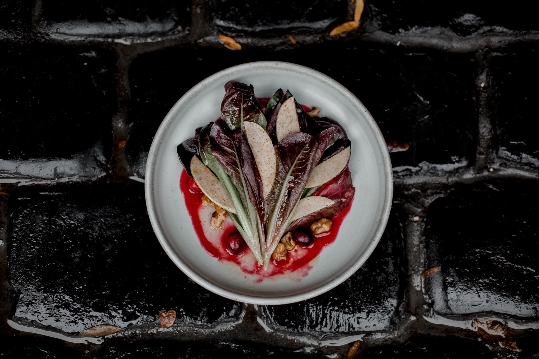 A top-down view of a plated salad on a wet dark cobblestone surface. The salad consists of red and purple radicchio leaves, sliced pears, walnuts, and a red sauce in a white plate.