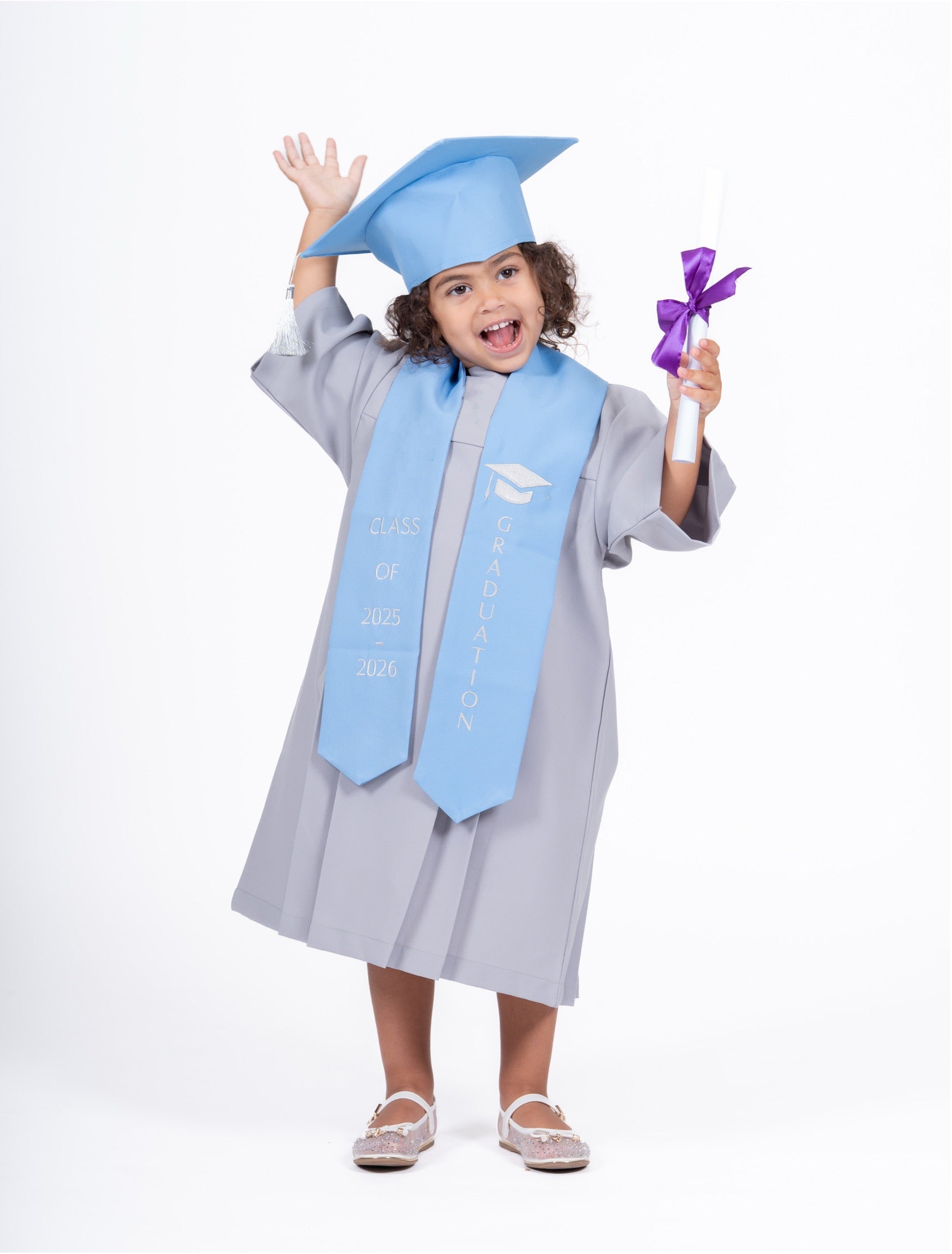 A young child wearing a light blue graduation cap, matching graduation gown, and a light blue satin sash with white text, holding a diploma scroll with a purple ribbon against a plain white studio background.